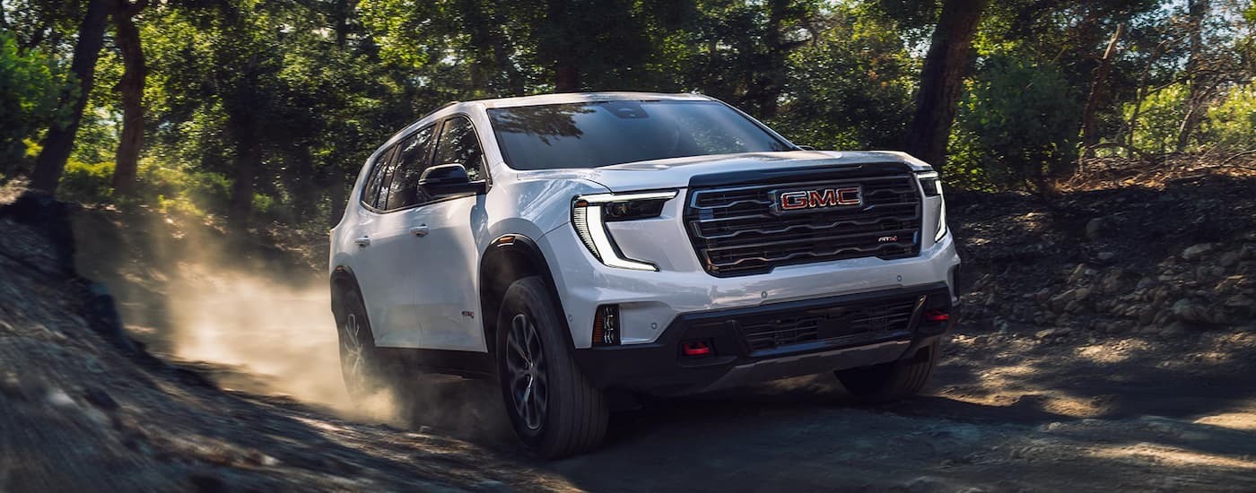 White 2026 GMC Acadia driving off-road in a forest.
