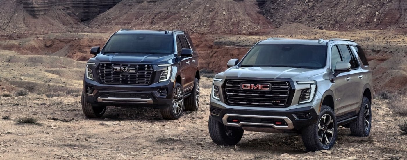 A black 2025 GMC Yukon Denali and a silver Yukon AT4 are parked off-road.